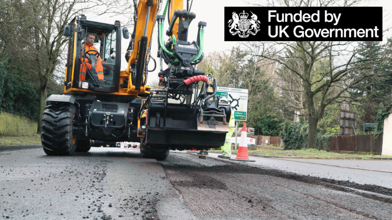 Image shows a road resurfacing machine with the words Funded by UK Government and their logo.