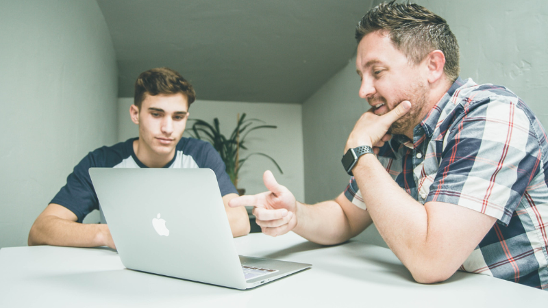 Man and teenage boy sat together looking at a laptop screen.