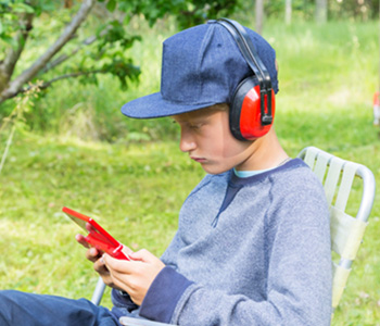 A child sitting outside on a chair wearing ear defenders. The child is playing on a gaming console.