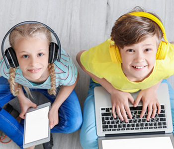Two children sitting on the floor wearing headphones looking up and smiling to the camera above.  The young girl holds an ipad and the boy is on a laptop.