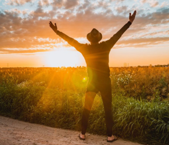 Photo of man stood outside with both arms held up while facing a bright sunset and overlooking a field.
