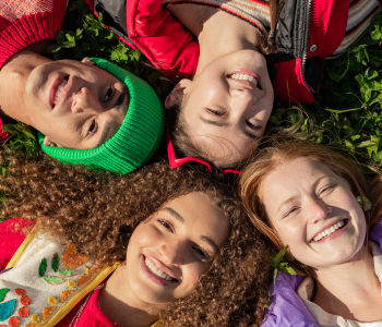 Photo of 4 teens laid down on the grass in a circle while looking upwards while smiling.