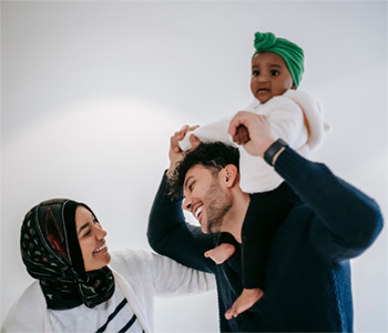 A young mother and father standing together. The father is carrying their young daughter on his shoulders. Both parents are looking at each other and smiling warmly.