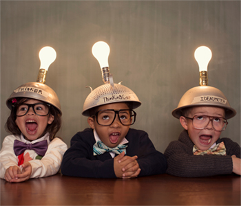 Three young children sitting side by side at a table. Each child has a metal colander with a light bulb on their head with a sticker. The different stickers read &#039;Thinker&#039;, &#039;Thinking Cap&#039; and &#039;Ideaometer&#039;.