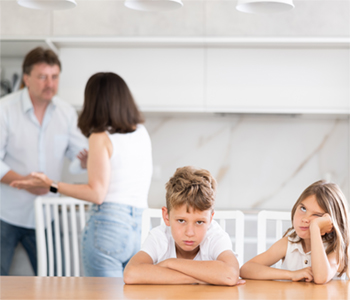 A young boy and a young girl sitting at a kitchen table. Both children look fed up. In the background, their mother and father are having an argument, seemingly unaware of the impact it is having on their children.