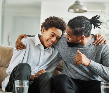 A young teenage boy sitting on a sofa, leaning towards his father. The father is looking at him with much love, and the boy is smiling warmly at his father. The father has his arm around the boy.